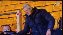 a man in a blue jacket stands in front of a stadium with a sky sports logo on the bottom right