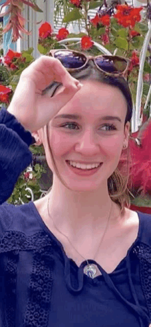 a young woman wearing sunglasses and a necklace is smiling in front of flowers .