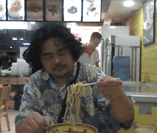 a man in a hawaiian shirt is eating noodles with chopsticks in a restaurant