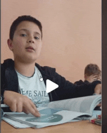 a boy wearing a tee sail shirt is sitting at a desk reading a book