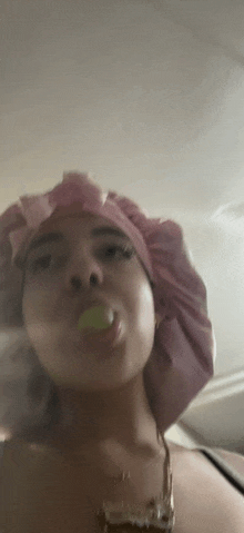 a woman wearing a pink shower cap is blowing a bubble with a green object in her mouth .