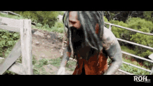 a man with dreadlocks and a beard is standing next to a wooden fence with the word roh written on the bottom