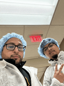 a red exit sign hangs from the ceiling above two men wearing scrub caps