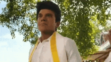 a man in a white suit and yellow sash is standing under a tree .