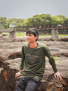 a young man wearing a green prada sweater sits on a rock