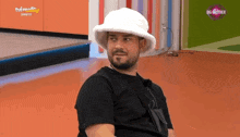 a man with a beard wearing a white hat and a black shirt is sitting on the floor .