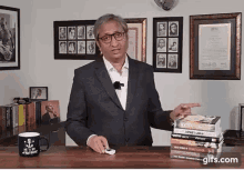 a man in a suit and glasses is standing in front of a table with a stack of books .