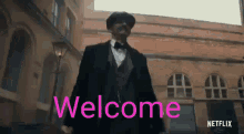 a man in a suit and bow tie is standing in front of a brick building with the words welcome written on it .