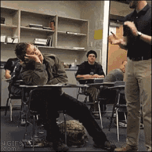 a man sitting at a desk in a classroom with his hand on his face while another man talks to him