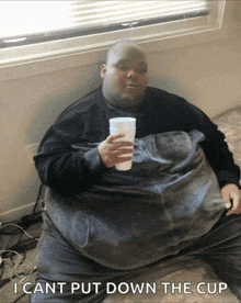 a very fat man is sitting on a bed holding a cup with the caption i cant put down the cup