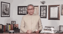 a man in a suit and glasses is standing in front of a stack of books and a framed picture of mahatma gandhi