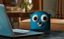 a blue stuffed animal with big eyes sits on a desk next to a laptop