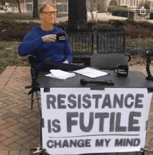 bill gates sits at a table with a sign that says resistance is futile change my mind