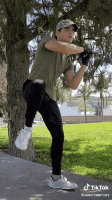 a young man wearing a hat and gloves is standing next to a tree in a park with a tiktok watermark