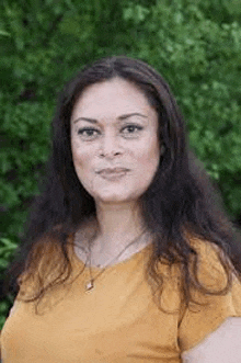 a woman in a yellow shirt and necklace is standing in front of a tree .