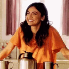 a woman in an orange shirt is smiling while sitting at a table with coffee cups and a coffee pot .