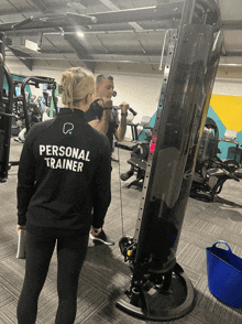 a woman wearing a personal trainer jacket watches a man lift weights in a gym