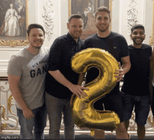 a man wearing a new haven gang shirt holds a large gold number 2 balloon