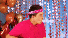 a man wearing a pink shirt and a bandana is blowing a bubble in front of balloons and stars .