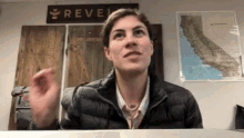 a woman in a black jacket is sitting at a desk in front of a wall with a map of california on it .