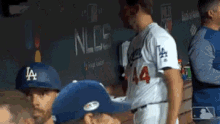 a group of baseball players are standing in a dugout . one of the players is wearing a la hat .