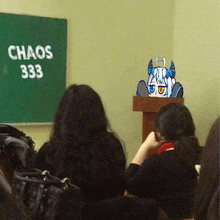 a girl with horns sits at a podium in front of a chalkboard that says chaos 333