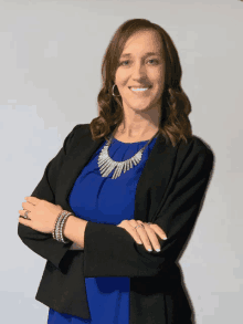 a woman in a blue shirt and black jacket stands with her arms crossed and smiles