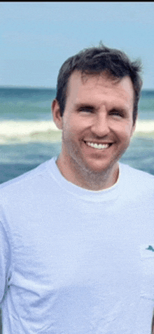 a man wearing a white t-shirt is smiling in front of the ocean