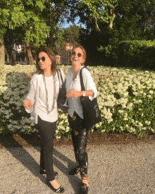 two women standing next to each other holding wine glasses in front of a bush with white flowers