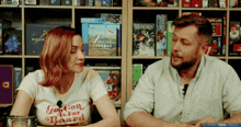 a man and a woman are sitting in front of a shelf full of board games including munchkin and 7 moons