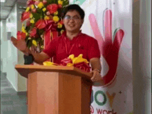 a man in a red shirt is standing at a podium with flowers in front of a sign that says joy work