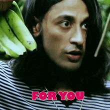 a man with long hair is holding a bunch of green bananas in front of a sign that says " for you "