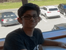 a young man wearing glasses is sitting at a table in front of a window with cars parked in the background .