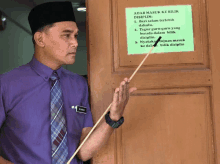 a man in a purple shirt and tie points to a sign that says " adar masuk ke bilik "