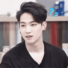 a young man wearing a black shirt and earrings is sitting in front of a bookshelf and looking at the camera .