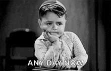 a black and white photo of a boy sitting at a desk with his hand on his chin and the words `` any day now '' .