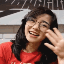 a woman wearing glasses and a red shirt is smiling and waving her hand