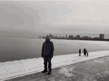 a man is standing on a frozen beach next to a body of water with a city skyline in the background .