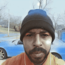 a man with a beard wearing a beanie looks at the camera with a blue car in the background