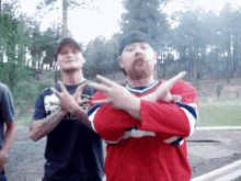 a man in a red shirt is smoking a cigarette while another man stands behind him with his arms crossed