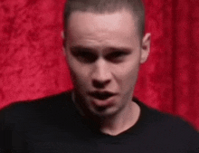 a man in a black shirt is standing in front of a red curtain and looking at the camera .