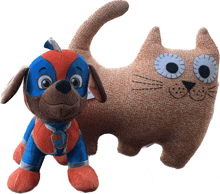 a stuffed dog in a superhero costume stands next to an orange cat