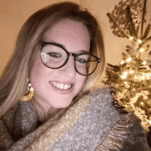 a woman wearing glasses and a scarf stands in front of a christmas tree