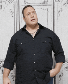 a man wearing a black shirt and a necklace is standing in front of a white wall