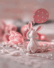 a white bunny holding a pink polka dot balloon is sitting on a pink surface