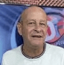 a bald man wearing a white shirt and a black necklace is smiling in front of a blue wall .