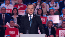 a man in a suit and tie stands at a podium in front of a crowd with signs that say duda 2020