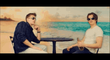 two men are sitting at a table in front of a beach