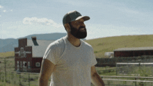 a man with a beard wearing a white shirt and a hat with the letter g on it stands in front of a barn