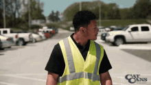 a man in a yellow vest is standing in a parking lot with a cat cans logo in the background